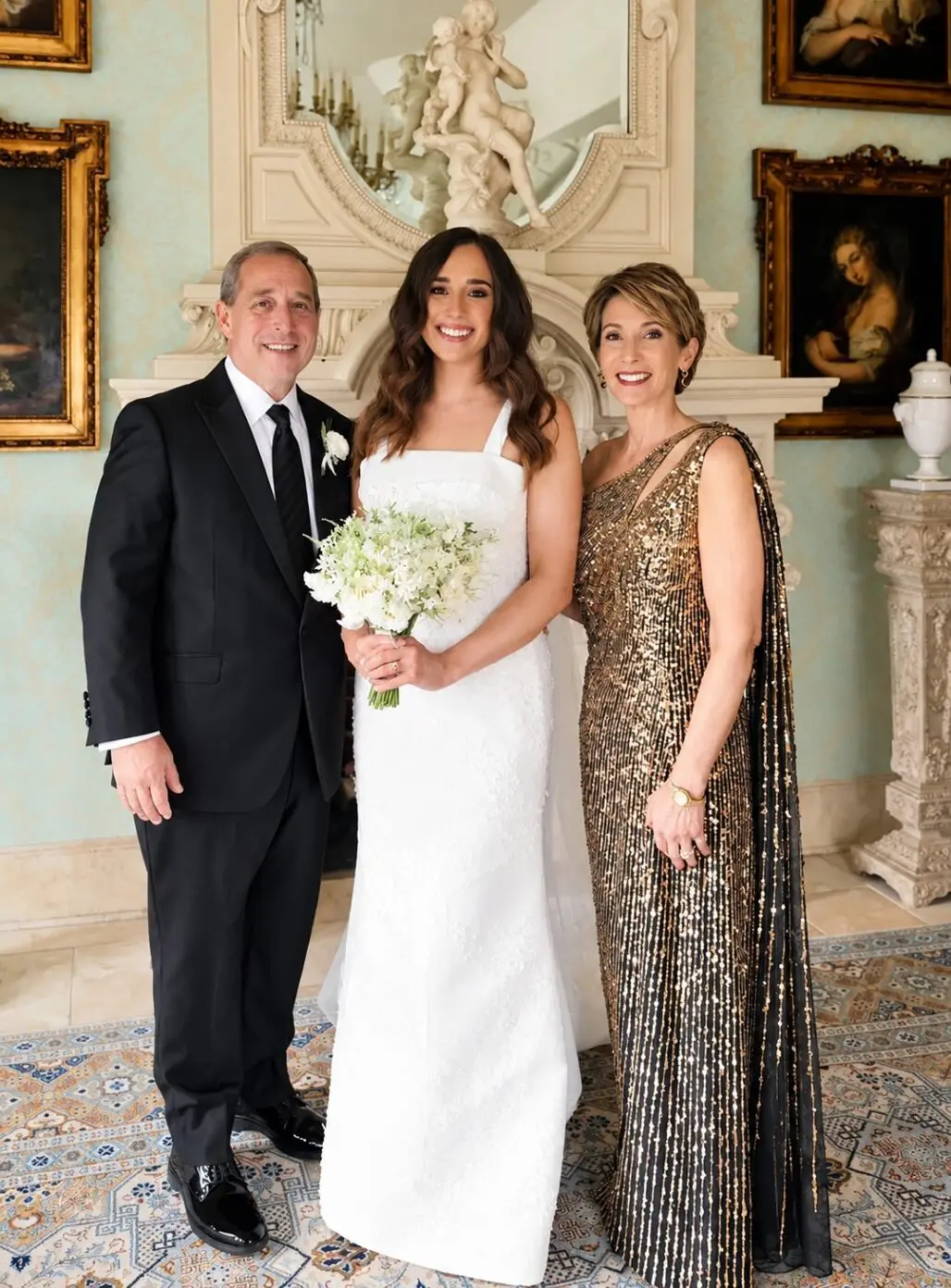 Mother of the bride in a gold sequined gown with a sheer black cape, standing beside the bride and the father of the bride